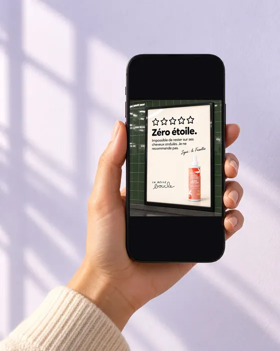 Une main tient un téléphone qui affiche la pub « Zéro étoile » de la Brume Anti-Frisottis de La Belle Boucle